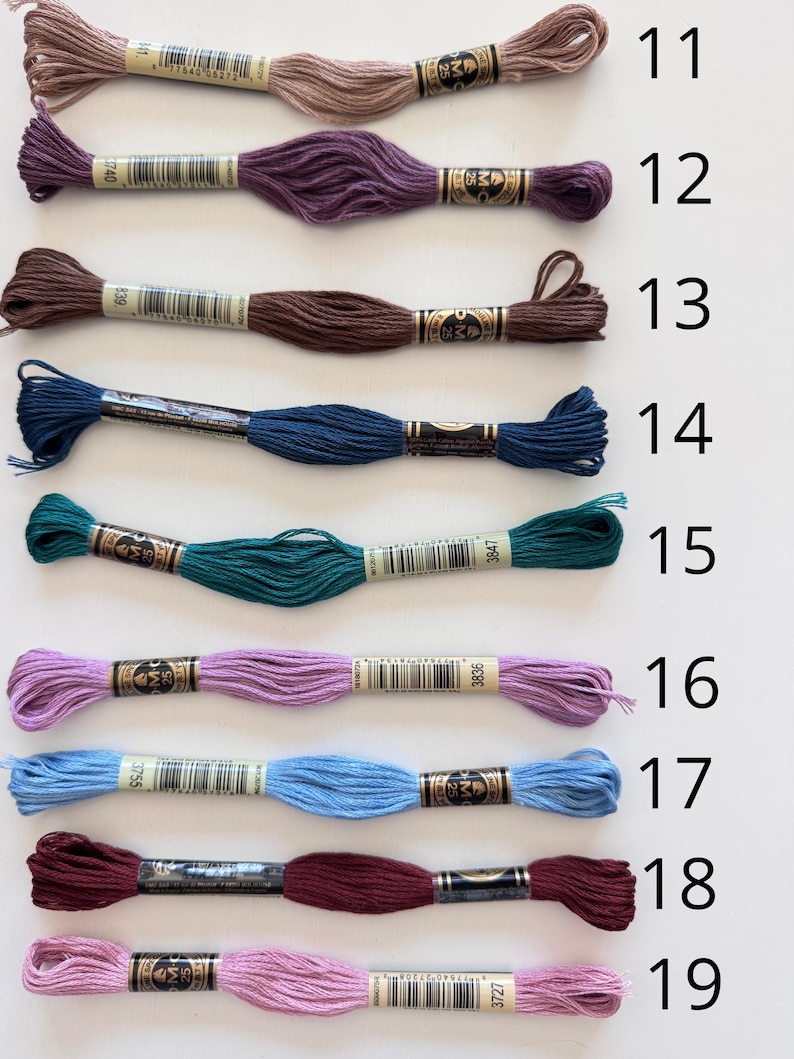 May include: Nine skeins of embroidery floss in various colors, including shades of brown, purple, teal, blue, and pink. Each skein is labeled with a number from 11 to 19. The floss is neatly arranged, showcasing the range of colors available.