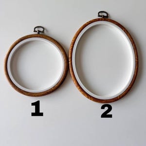 May include: Two embroidery hoops, one round and one oval, with light brown wooden frames and white interiors. The hoops have a metal clasp at the top. The number 1 is below the round hoop, and the number 2 is below the oval hoop.