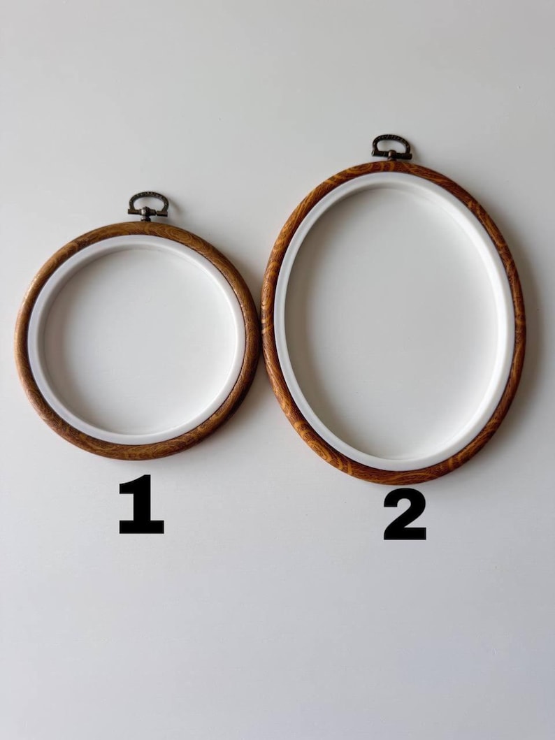 May include: Two embroidery hoops, one round and one oval, with light brown wooden frames and white interiors. The hoops have a metal clasp at the top. The number 1 is below the round hoop, and the number 2 is below the oval hoop.