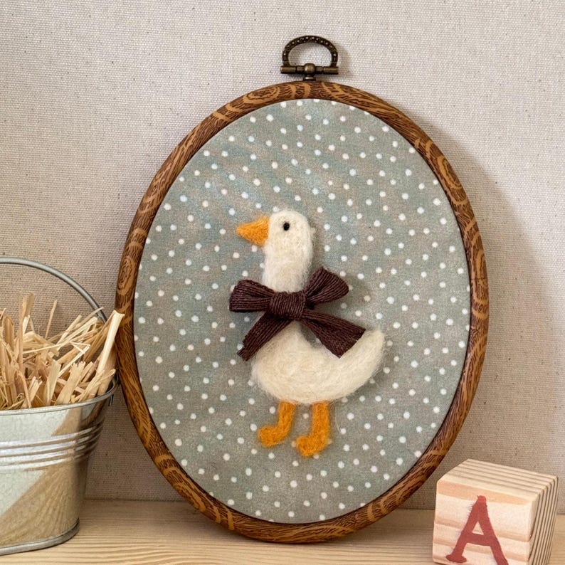 May include: Oval-shaped wall hanging featuring a felt goose with a brown bow, orange beak and feet, set against a gray polka-dot background. The goose is framed by a wooden hoop with a metal loop for hanging.