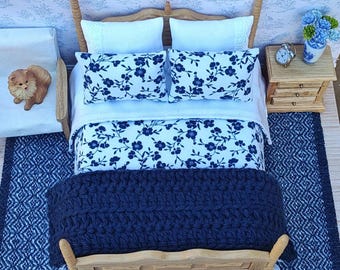 Puede incluir: Escena de dormitorio en miniatura con una cama de madera, ropa de cama blanca y almohadas con estampados florales azules. Una manta de punto azul marino cubre los pies de la cama. Una pequeña mesita de noche de madera con un reloj y un jarrón de flores. Una alfombra y una silla en miniatura completan la escena.