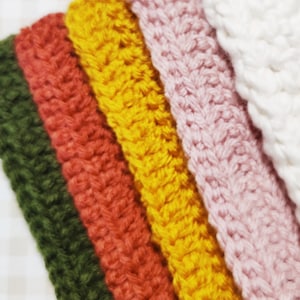 May include: Five crocheted headbands in different colors: dark green, red, yellow, pink, and white. The headbands are made with a simple stitch pattern.