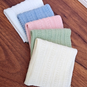 Op de afbeelding: Vijf gevouwen gebreide stoffen in wit, lichtblauw, roze, groen en crème. Elk stuk heeft een textuur, openwerk ontwerp. De stoffen zijn diagonaal op een houten oppervlak gerangschikt, waarbij hun kleuren en texturen worden getoond.