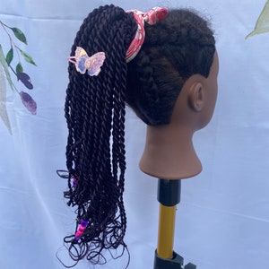 May include: A black braided ponytail with pink and purple beads, a pink and white patterned hair tie, and a butterfly hair clip.