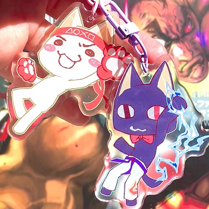 May include: Two clear acrylic keychains featuring cartoon characters. The first keychain features a white cat with red boxing gloves and a red headband with the text "DOXO". The second keychain features a purple cat with a blue karate belt and a red bow tie.