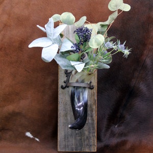 May include: A rustic wall-mounted vase made from a polished horn, mounted on a weathered wooden plank. The horn vase is filled with a variety of faux greenery and flowers, including silver-leafed plants and blue thistle-like blooms. The vase is held in place by a decorative metal bracket.