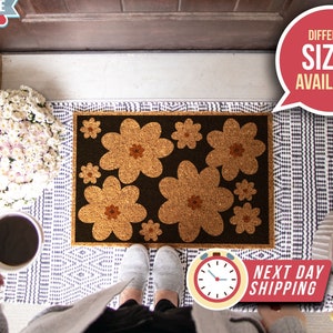 Op de afbeelding: Een bruine kokosvezel deurmat met een zwarte achtergrond en een ontwerp van bruine bloemen. De deurmat ligt op een wit en blauw gepatroneerd kleed. De tekst "Next Day Shipping" is zichtbaar in de rechterbenedenhoek van de afbeelding.