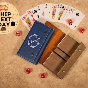 May include: A blue and brown leather card and dice case with the initials "K.M." engraved on the front. The case is open and has three slots for cards and dice. There are playing cards and dice scattered around the case.