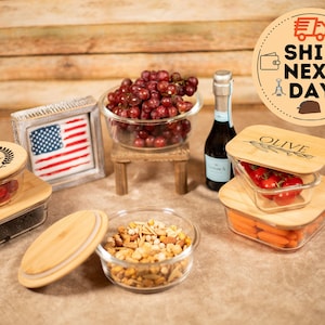 May include: Set of six glass food storage containers with bamboo lids. The lids are engraved with various designs, including a monogram, an olive branch, and the word "OLIVE".