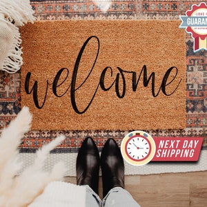May include: A brown coir doormat with the word "welcome" written in black cursive script. The doormat is on a patterned rug with a white blanket and a pair of black shoes on top of the rug.