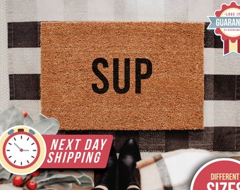May include: A brown coir doormat with the word "SUP" in black lettering. The doormat is on a black and white checkered rug.