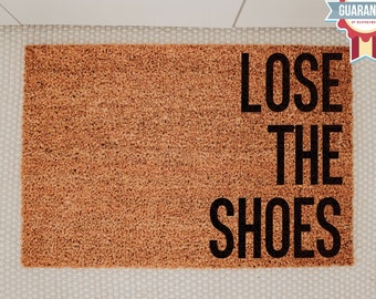 May include: A brown coir doormat with the text "Lose the shoes" in black lettering.