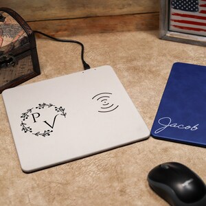 May include: Two wireless charging pads, one white with a floral monogram "PV" and a wireless charging symbol, and one blue with the name "Jacob" in white script. A black computer mouse is in the foreground.