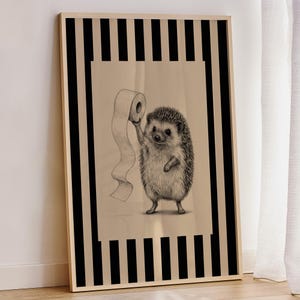 May include: A framed wall art print featuring a detailed pencil-style illustration of a hedgehog holding a roll of toilet paper. The central image is set against a beige background, framed by a bold border of vertical black and beige stripes.