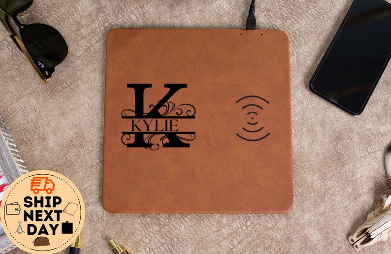 May include: A brown leather wireless charging pad with a black monogrammed initial "K" and the name "KYLIE" engraved on the left side. The charging pad has a wireless charging symbol on the right side.