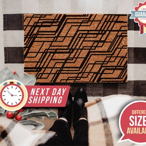 May include: A brown doormat with a black geometric pattern. The doormat is made of coir and has a textured surface. The text "Next Day Shipping" is printed on a red banner below the doormat.