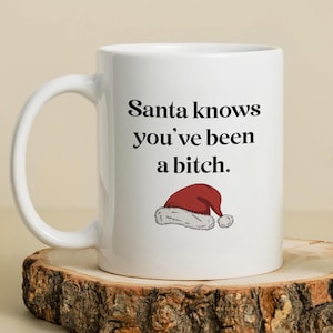 May include: White ceramic mug with the text "Santa knows you've been a bitch." and a Santa hat illustration. The mug is on a wooden surface, perfect for a holiday gift or a humorous addition to a kitchen.
