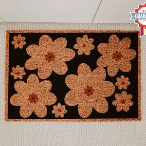 Op de afbeelding: Een bruine deurmat met een zwarte achtergrond en een patroon van oranje bloemen. De bloemen zijn gestileerd en hebben een retro-look.