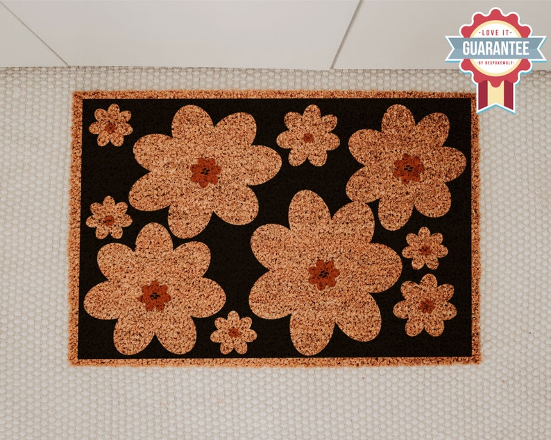 Op de afbeelding: Een bruine deurmat met een zwarte achtergrond en een patroon van oranje bloemen. De bloemen zijn gestileerd en hebben een retro-look.