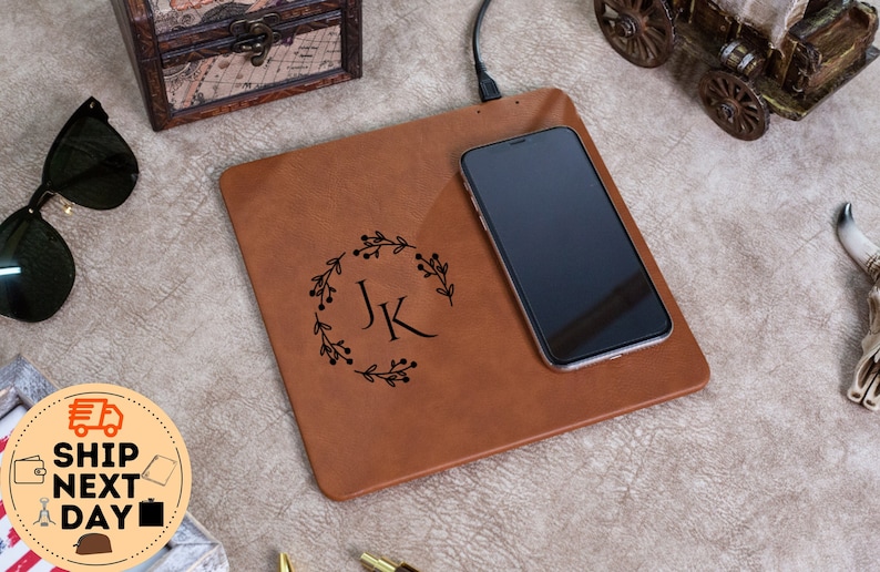 May include: A brown leather wireless charging pad with a phone on top. The pad has a monogram of "JK" in a wreath design.