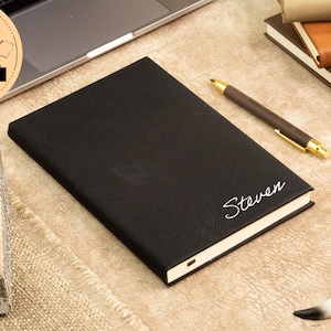 May include: Black leather-bound journal with the name "Steven" embossed in white on the front cover. The journal is lying on a beige surface with a pen and a decorative skull nearby.