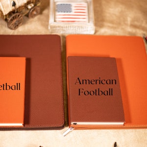 May include: Two brown leather journals with orange covers. The smaller journal has the text "Basketball" on the front. The larger journal has the text "American Football" on the front.