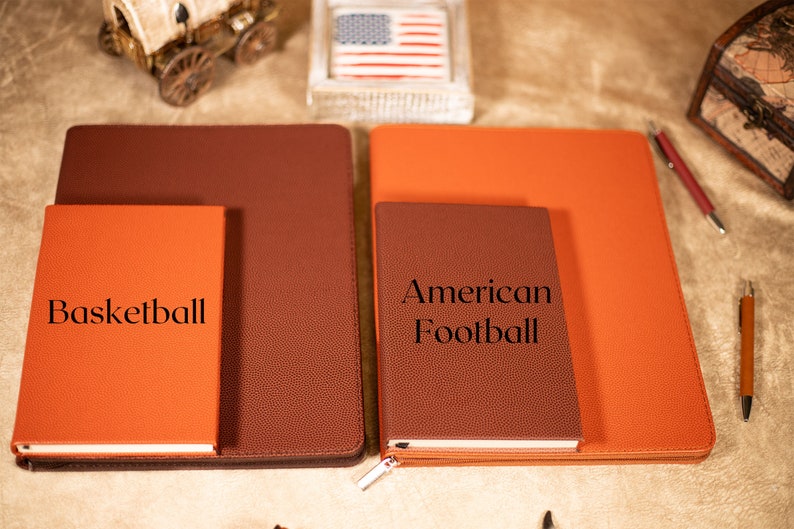 May include: Two brown leather journals with orange covers. The smaller journal has the text "Basketball" on the front. The larger journal has the text "American Football" on the front.
