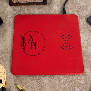 May include: Red leather wireless charging pad with a monogrammed design in a circle with a leaf design and the initials "AH". The charging pad has a wireless charging symbol on the right side.