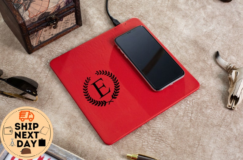 May include: A red leather wireless charging pad with a black engraved monogram of the letter "E" surrounded by a laurel wreath. A black smartphone is resting on the charging pad.
