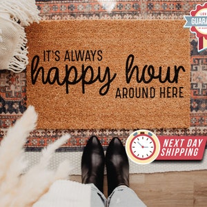 May include: A brown coir doormat with the text "It's always happy hour around here" in black lettering. The doormat is on a patterned rug with a white throw blanket and a bouquet of dried flowers.