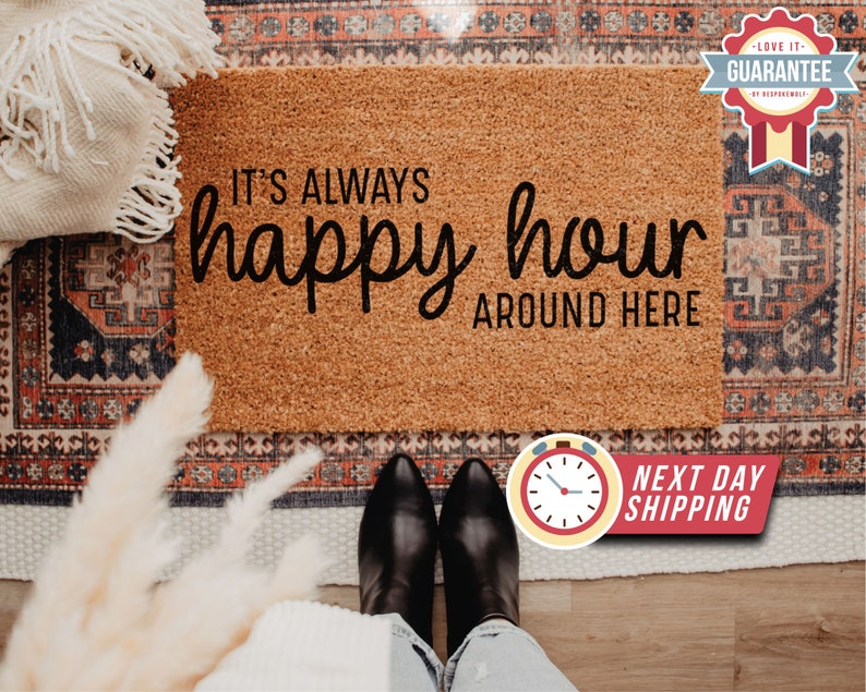 May include: A brown coir doormat with the text "It's always happy hour around here" in black lettering. The doormat is on a patterned rug with a white throw blanket and a bouquet of dried flowers.