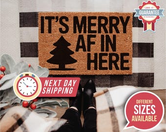May include: A rectangular doormat with the text "IT'S MERRY AF IN HERE" and a black Christmas tree graphic. The mat is brown with black lettering and a black tree. The mat is on a black and white checkered background.