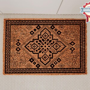 K&ouml;nnte beinhalten: Eine braune Kokosmatte mit einem schwarz-wei&szlig;en geometrischen Muster. Das Muster zeigt eine zentrale Diamantform mit komplizierten floralen Designs.