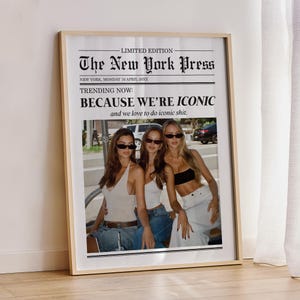May include: A framed print featuring a newspaper-style design with the headline "The New York Press." The text reads "TRENDING NOW: BECAUSE WE'RE ICONIC." Below the text is a photograph of three women wearing sunglasses. The frame is a light wood color.