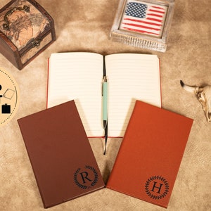 May include: Two brown leather journals with personalized initials, "R" and "H", in a wreath design. The journals are open to blank pages with a blue pen on top of one of the pages.