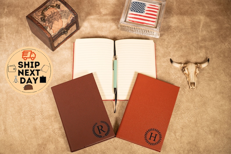 May include: Two brown leather journals with personalized initials, "R" and "H", in a wreath design. The journals are open to blank pages with a blue pen on top of one of the pages.