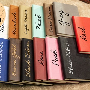 May include: A variety of leather-bound journals in different colors, including blue, brown, black, red, teal, pink, gray, and rawhide. Each journal has a different color and a gold or white inscription on the front cover. The inscriptions include "Blue Silver", "Brown Gold", "Black Silver", "Rose", "Pink", "Black Silver", "Teal", "Dark Brown", "Red", "Grey", and "Rawhide".