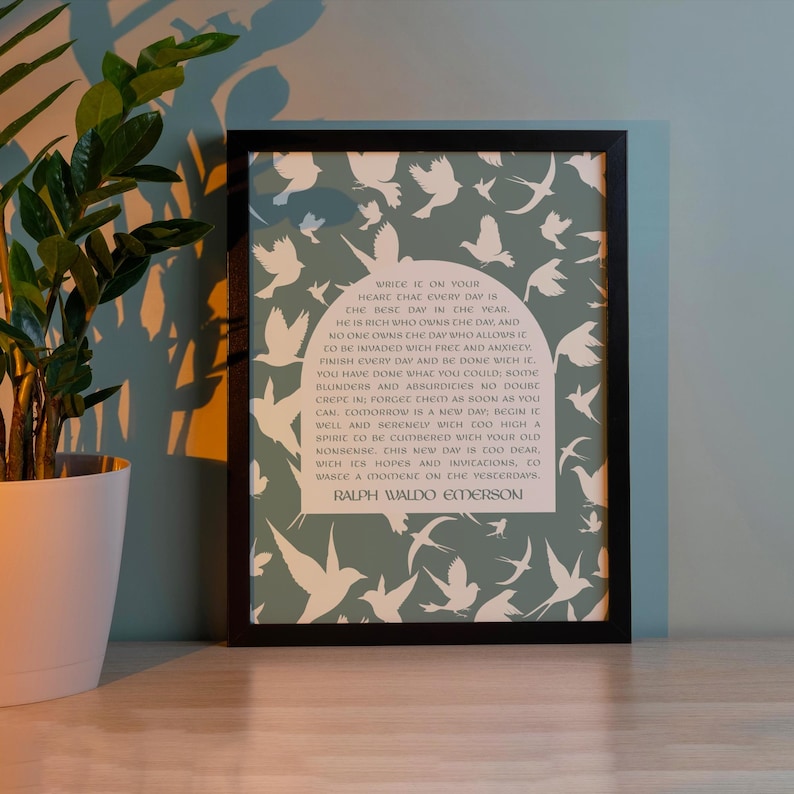 May include: Framed print with a quote by Ralph Waldo Emerson, set against a sage green backdrop with white dove silhouettes. The print is in a black frame. A potted plant is nearby.