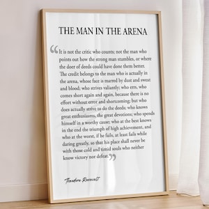 May include: A framed print featuring the quote "The Man in the Arena" by Theodore Roosevelt. The text is in a classic serif font on a white background, encased in a light wood frame. The quote is a call to action.