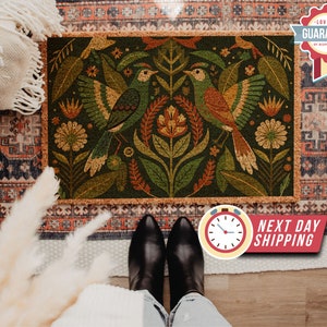 May include: A cork doormat with a green background featuring a stylised floral design with two birds in orange, yellow, and brown. The birds are facing each other and have their wings spread out. The doormat has a brown border.