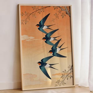 May include: Framed art print featuring five swallows in flight against an orange and yellow gradient background. The swallows have blue wings, white bellies, and red faces. Branches with leaves are in the corners of the print.