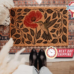 Op de afbeelding: Rechthoekige deurmat met een kolibrie en rood bloemmotief. De kolibrie wordt in vlucht afgebeeld, met zijn snavel uitgestrekt naar de bloem. De mat heeft een natuurlijk, getextureerd uiterlijk met zwarte bladeren.