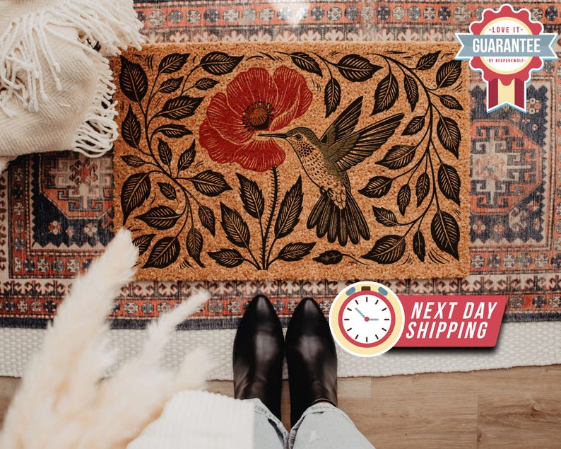 Op de afbeelding: Rechthoekige deurmat met een kolibrie en rood bloemmotief. De kolibrie wordt in vlucht afgebeeld, met zijn snavel uitgestrekt naar de bloem. De mat heeft een natuurlijk, getextureerd uiterlijk met zwarte bladeren.