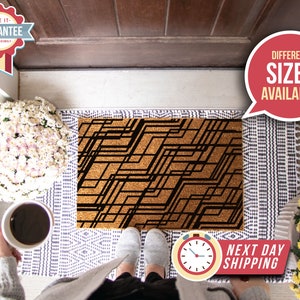 May include: A brown coir doormat with a black geometric pattern. The doormat is placed on a white and gray patterned rug. The text "Different Sizes Available" is in a red speech bubble in the upper right corner of the image.