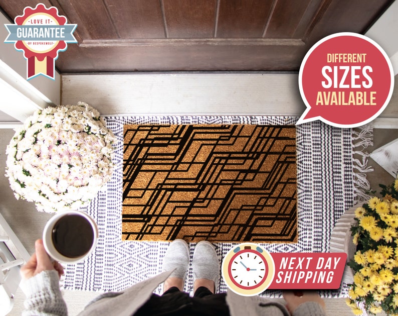 May include: A brown coir doormat with a black geometric pattern. The doormat is placed on a white and gray patterned rug. The text "Different Sizes Available" is in a red speech bubble in the upper right corner of the image.