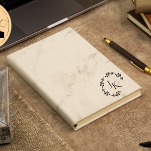May include: A white marble notebook with a black floral wreath and the initials "JK" in the center. The notebook is lying on a brown surface with a pen and a decorative skull.