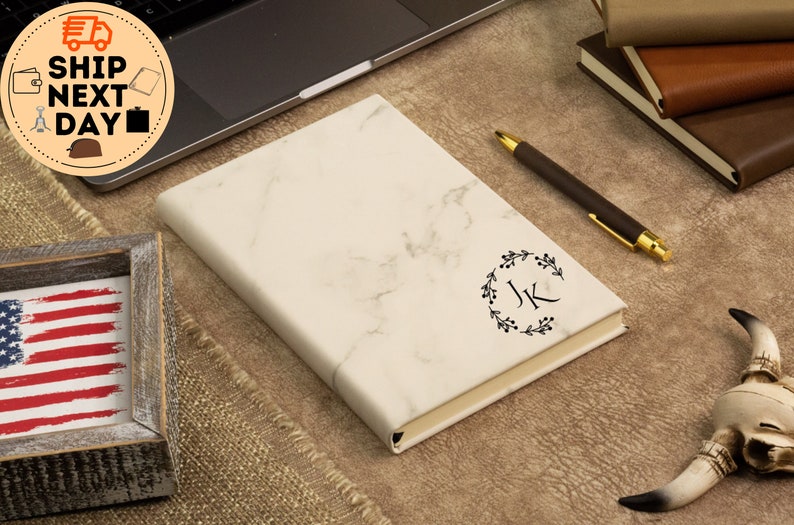 May include: A white marble notebook with a black floral wreath and the initials "JK" in the center. The notebook is lying on a brown surface with a pen and a decorative skull.