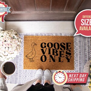 May include: A rectangular coir doormat with a goose illustration and the text "GOOSE VIBES ONLY" in black. The mat sits on a patterned rug. The image includes a person's feet, a coffee cup, and a sign indicating different sizes are available.