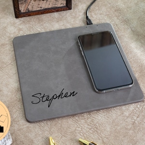 May include: A grey leather wireless charging pad with the name "Stephen" engraved on it. A black smartphone is charging on the pad.