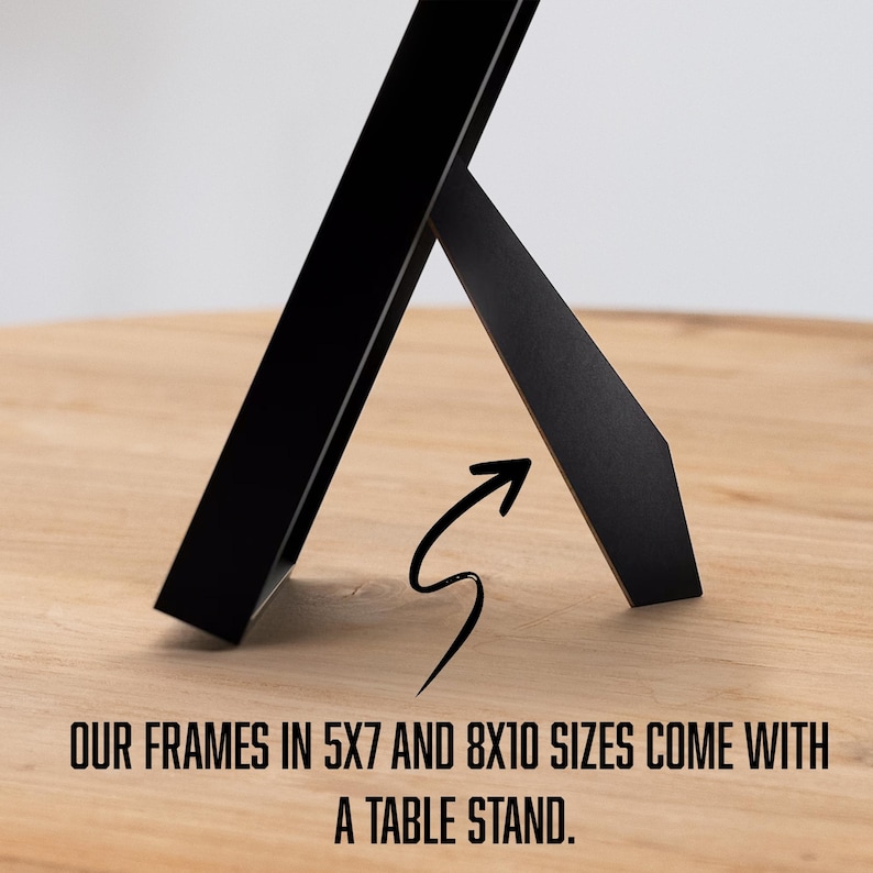 May include: Black picture frame with a table stand, on a light wood surface. The frame is angled, showing its design. Text at the bottom reads: "OUR FRAMES IN 5X7 AND 8X10 SIZES COME WITH A TABLE STAND."